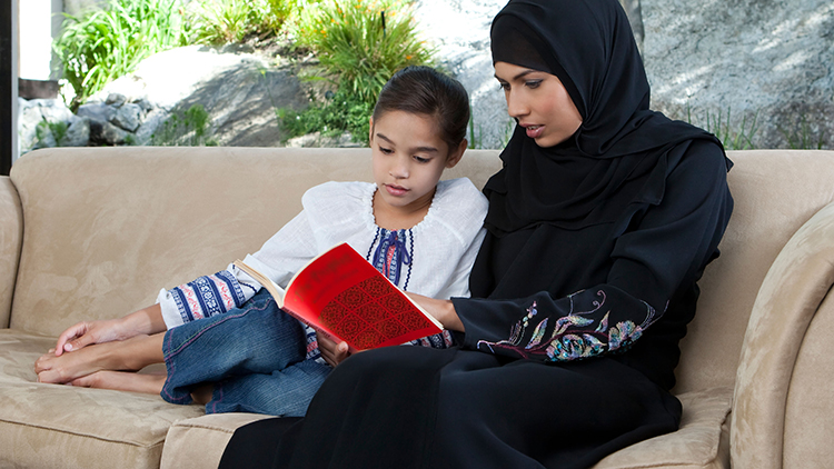 A young Arab woman, wearing a traditional black hijab, sitting next to her 8-9 year old daughter and reading a book together